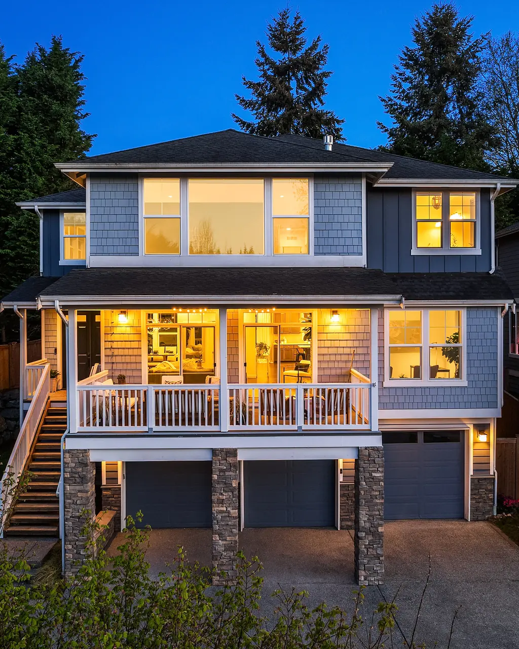 Two-story gray shingle home with a covered deck and three-car garage lit at twilight